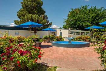 a fountain sits in the middle of a brick patio with colorful flowers and umbrellas